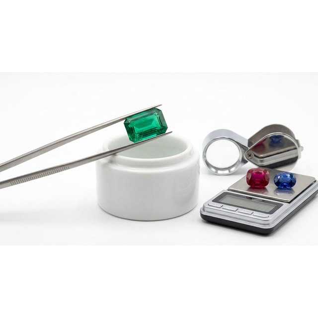 Tweezers holding an emerald-cut emerald with a loupe, scale and coloured stones